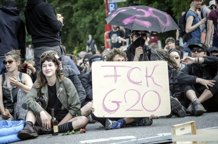 Demonstranten blockieren eine Straße und halten ein Schild "FCK G20" in den Händen. Foto: Tim Wagner