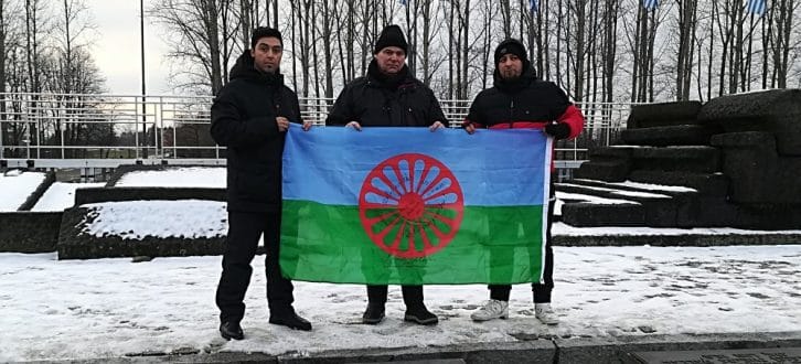 Richard Gauch (Mitte) zeigt mit zwei Begleitern die Flagge der Roma vor den ehemaligen Krematorien von Auschwitz-Birkenau. Foto: Ricky Burzlaff