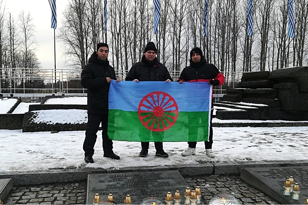 Richard Gauch (Mitte) zeigt mit zwei Begleitern die Flagge der Roma vor den ehemaligen Krematorien von Auschwitz-Birkenau. Foto: Ricky Burzlaff