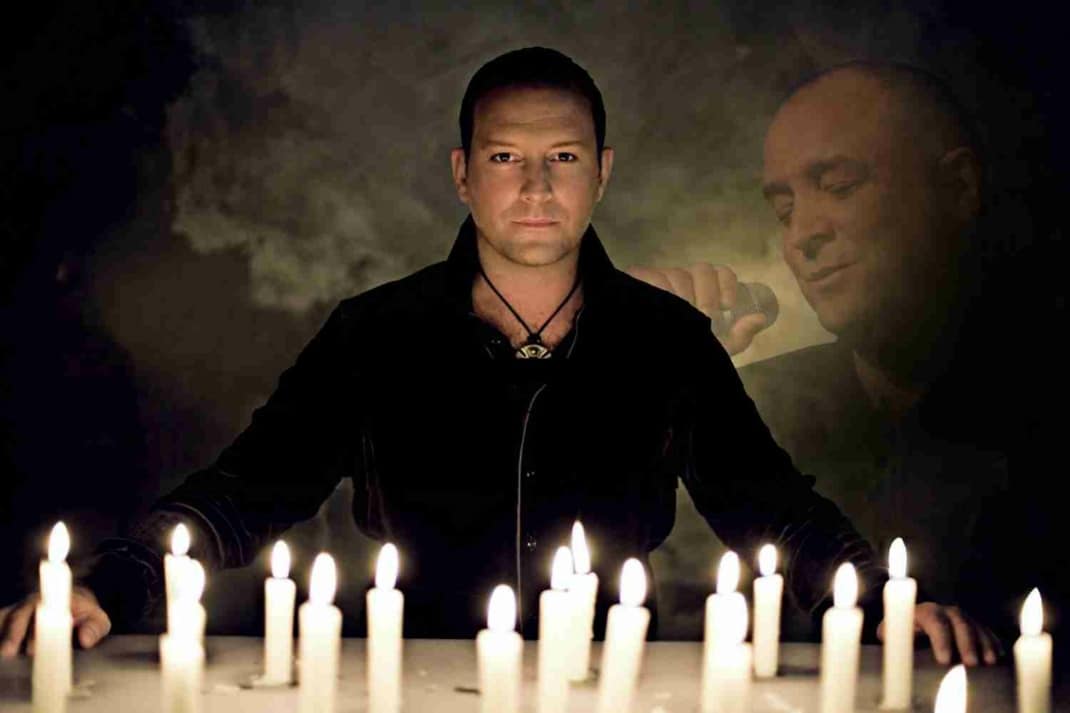 Eine Person steht mit leicht ausgebreiteten Armen vor einem Tisch, auf dem sehr viele brennende Kerzen stehen.