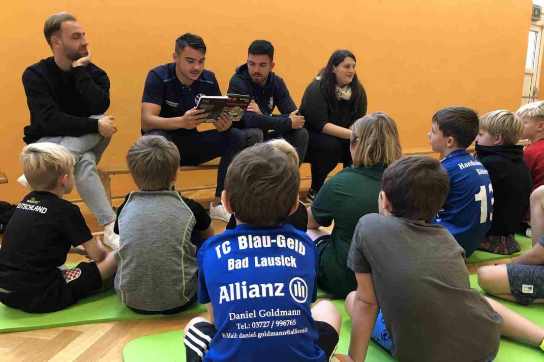 Fußballer von Lok Leipzig lesen Kindern etwas vor, die vor ihnen auf dem Boden sitzen