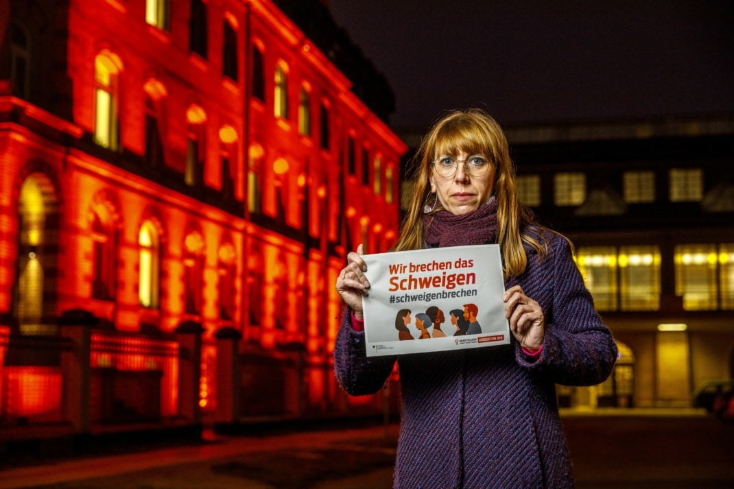 Gleichstellungsministerin Katja Meier vor dem orange angestrahlten Gebäude des Justiz- und Gleichstellungsministeriums am „World Orange Day“ 2021 mit einem Plakat in der Hand auf dem steht: Wir brechen das Scheigen, darunter der Hashtag #schweigenbrechen