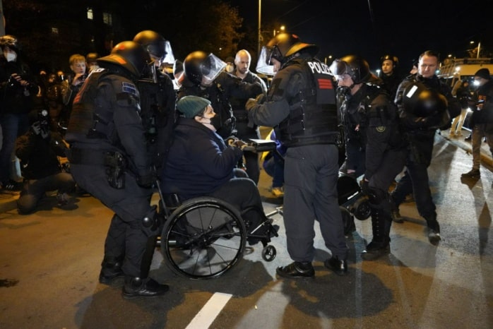 Eine Person im Rollstuhl wird von Polizist*innen weg getragen.