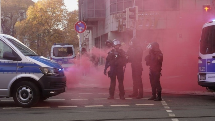 Pinker Rauch steht zwischen zwei Polizeiautos auf, dazwischen stehen mehrere Polizist*innen.