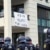 Von einem Balkon hängt ein Banner mit der Aufschrift "Kein Platz für Nazis". Davor läuft eine Gruppe Polizist*innen.