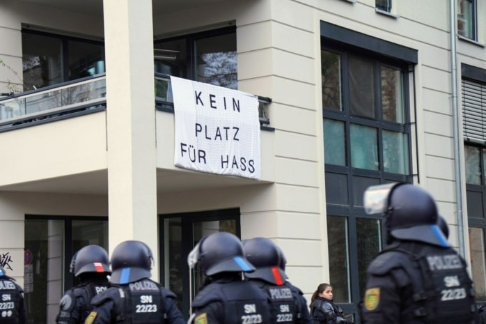 Von einem Balkon hängt ein Banner mit der Aufschrift "Kein Platz für Nazis". Davor läuft eine Gruppe Polizist*innen.
