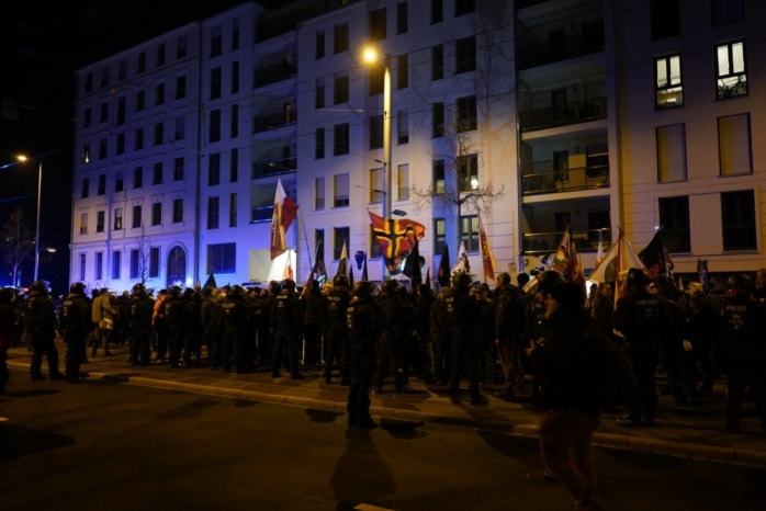 Hunderte Menschen stehen auf der Straße vor einer Gruppe Polizist*innen.