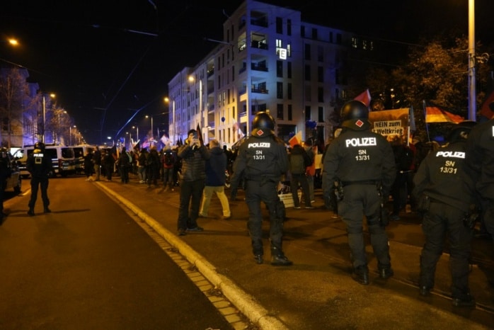 Hunderte Menschen stehen auf der Straße vor einer Gruppe Polizist*innen.
