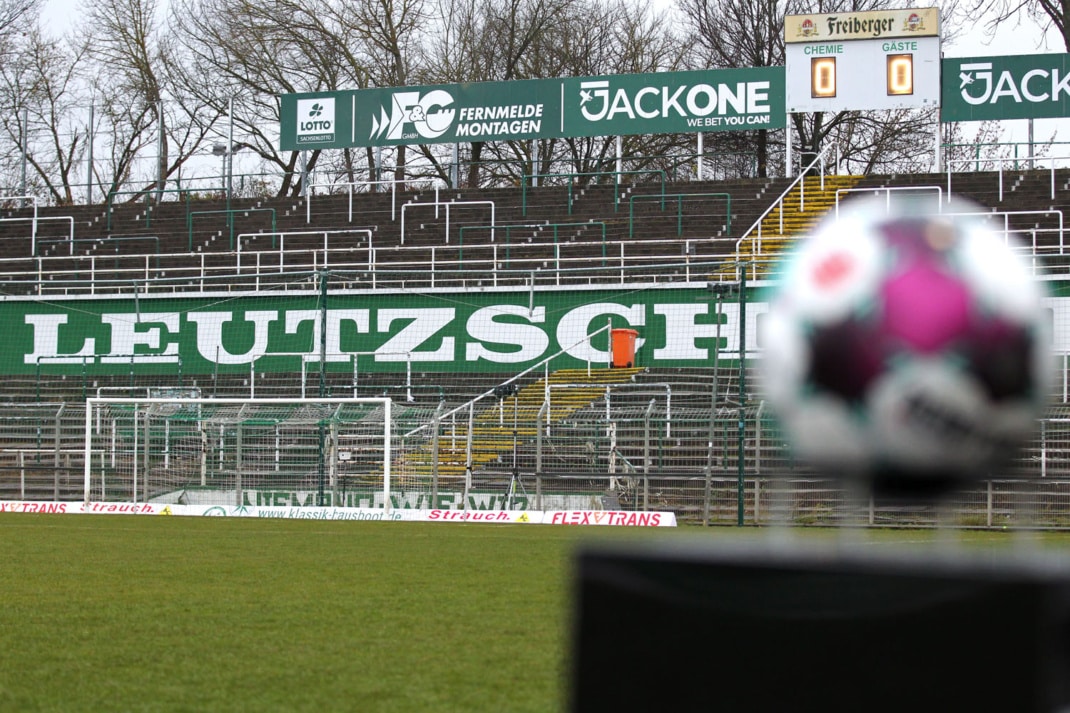 Blick auf den leeren Norddamm im Alfred-Kunze-Sportpark. Zu erkennen ist der Schriftzug "LEUTZSCH" in weiß auf grünem Untergrund. Daneben, im Vordergrund, ist verschwommen ein Fußball zu sehen. Foto: Jan Kaefer
