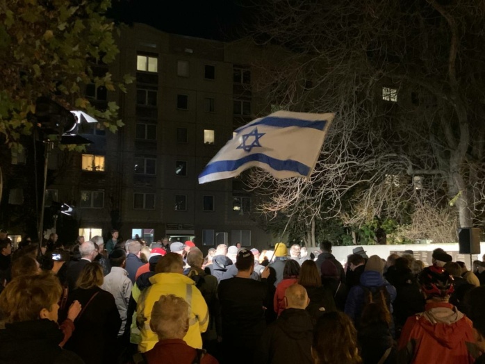 Auf einem Platz stehen mindestens hundert Personen vor dem Leipziger Synagogendenkmal in der Gottschedstraße. Die Männer tragen eine Kompfbedeckung, über den Personen wird eine Israelfahne.