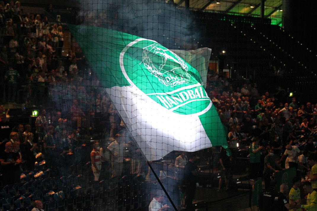 Eine große grün-weiße Fahne mit dem Vereinswappen des SC DHfK Handball wird in der abgedunkelten Halle von einem Lichtstrahl erleuchtet. Foto: Jan Kaefer