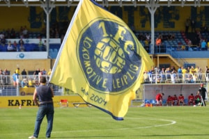 Eine Person steht auf dem Fußballplatz und hält eine große quadratische gelbe Fahne in der Hand, auf der das blaue Vereinswappen des 1. FC Lok Leipzig zu sehen. Foto: Jan Kaefer