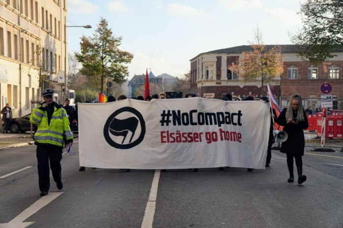 Eine Versammlung mit einem Banner der Aufschrift "No Compact, Elsässer go home" läuft auf einer Straße.
