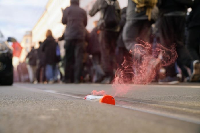 Auf einer Straße liegt leicht rauchende Pyrotechnik. Im Hintergrund laufen mehrere dutzen schwarz gegleideter Menschen.