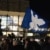 Eine blaue Fahne mit einer Friedenstaube weht über mehreren Personen auf einer Kundgebung.