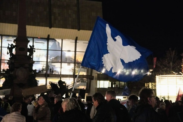 Eine blaue Fahne mit einer Friedenstaube weht über mehreren Personen auf einer Kundgebung.