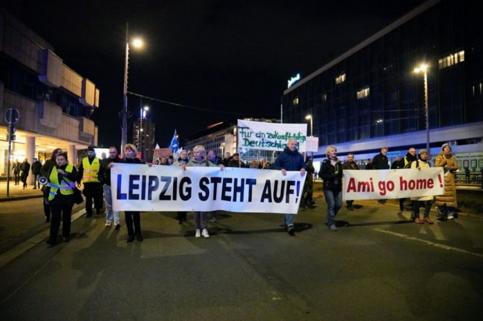 Personen mit Bannern laufen auf einer Straße. Auf den Bannern steht „Leipzig steht auf!“ und „Ami go home!“ geschrieben.