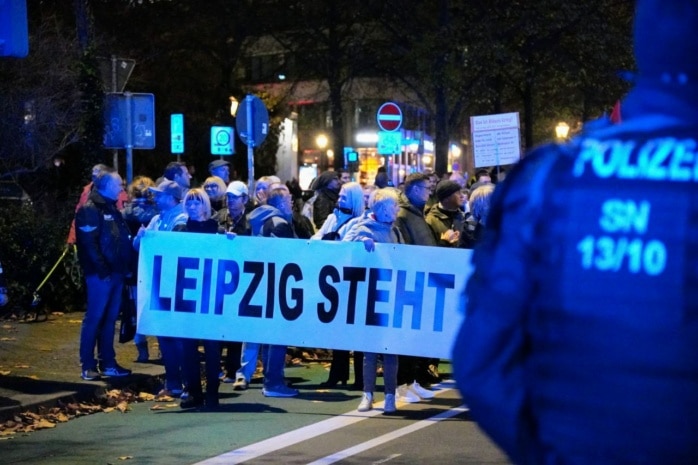 Menschen mit einem Banner, auf dem zu lesen ist „Leipzig steht ...“ stehen auf der Straße. im Vordergrund steht ein Polizeibeamter.
