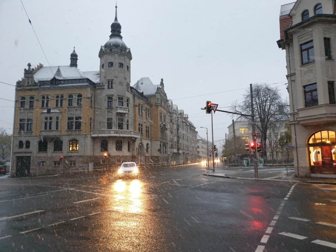 Ansicht einer Kreuzung, über die ein Auto mit Schweinwerfern fährt. Im Licht ist der Schneefall zu sehen.