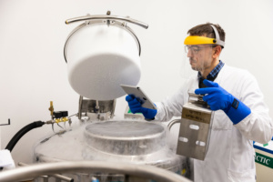 Dr. Vucinic in Schutzausrüstung vor einem sogenannten Kryotank und einer Kassette mit kryokonservierten, also tiefgefrorenen CAR-T-Zellen