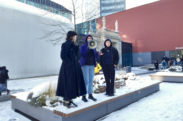 Drei Aktivist/-innen von „End Fossil“ informieren Studierende auf dem Campus Augustusplatz der Uni Leipzig am 13.12.2022 über die fortlaufende Besetzung des Audimax, sie stehen mit einem Megafon auf einer Sitzgelegenheit im Innenhof des Campus.