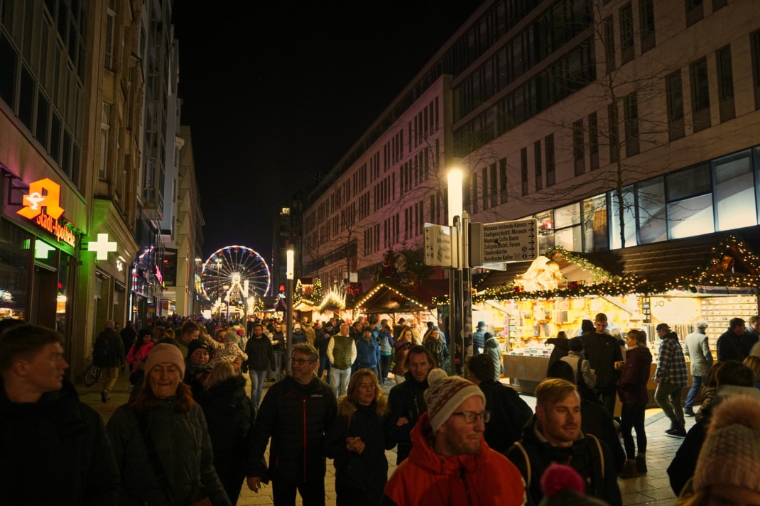 In der Grimmaischen Straße sind mehrere Stände des Weihnachtsmarktes zu sehen, im Hintergrund ein erleuchtetes Riesenrad. Dazwischen spazieren dutzende Menschen in zum Teil weihnachtlicher Kleidung.