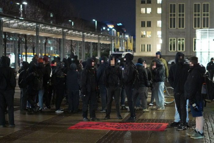 Personengruppe, größtenteils schwarz bekleidet, steht vor einem roten Banner