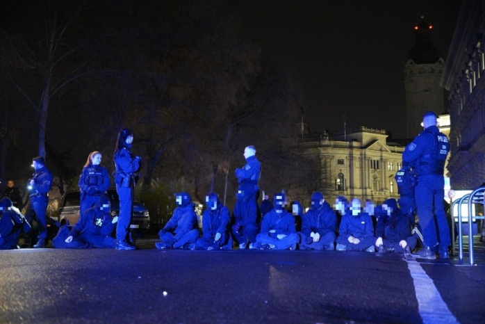 Schwarz gekleidete Personen sitzen auf der Straße, flankiert von Polizei