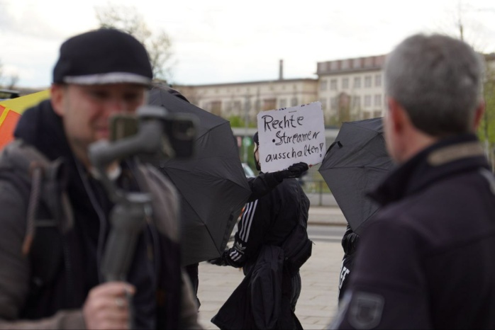 Person mit Handykamera im Vordergrund, im Hintergrund ein Banner und eine weitere Person