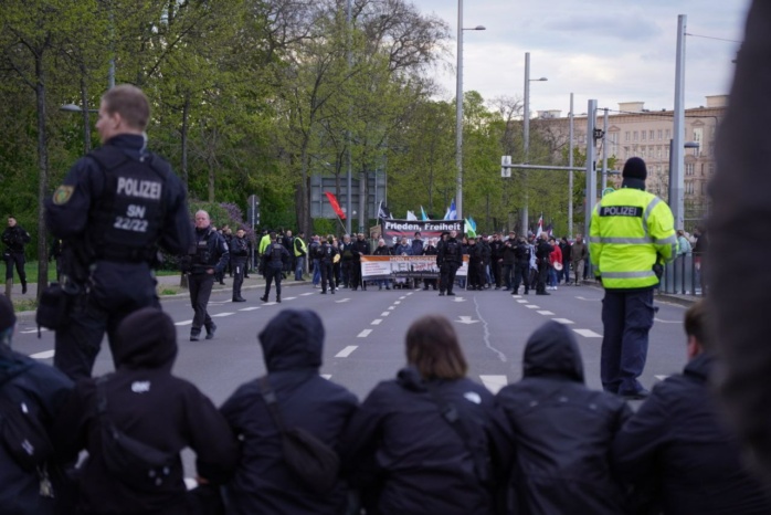 Mehrere schwarz gekleidete Personen, zu sehen von hinten, sitzen auf einer Straße. Im Vordergrund eine Gruppe, die auf der Straße demonstriert