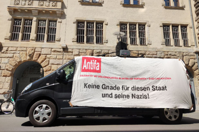 Personen hinter einem Banner verdeckt. Das Banner trägt die Aufschrift "Keine Gnade für diesen Staat und seine Nazis"