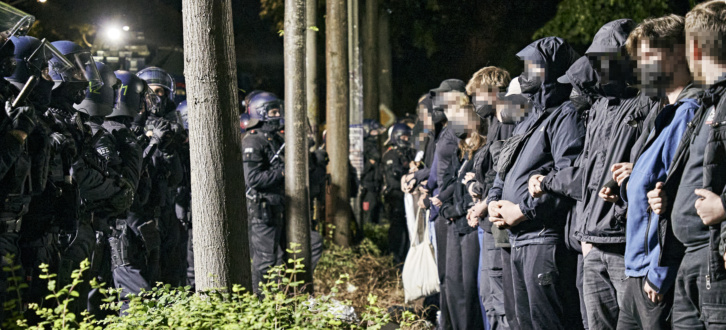 Dutzende Demonstrierende stehen einer Polizeikette gegenüber, die die Gruppe umschließt. die Demonstrierenden sind untereinander eingehakt, ihre Gesichter wurden unkenntlich gemacht.