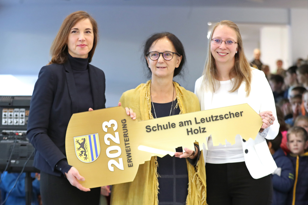 Drei Personen bei symbolischer Schlüsselübergabe.