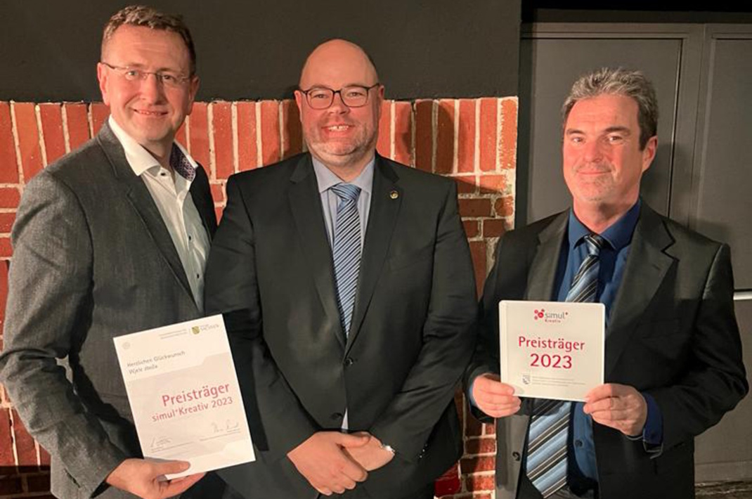 Drei Männer in Anzügen, zwei halten Schriftstücke.