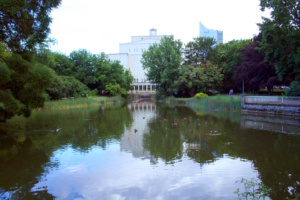 Parkanlage mit Teich, Gebäude im Hintergrund.