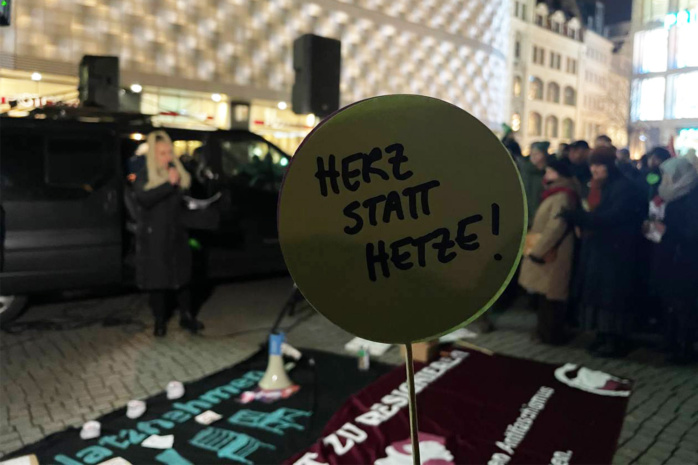 Im Vordergrund ein Schild, auf dem "Herz statt Hetze" steht. Im Hintergrund Menschen