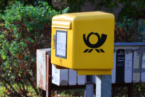 Gelber Briefkasten seitlich mit Postsymbol, an einem Aufsteller befestigt.