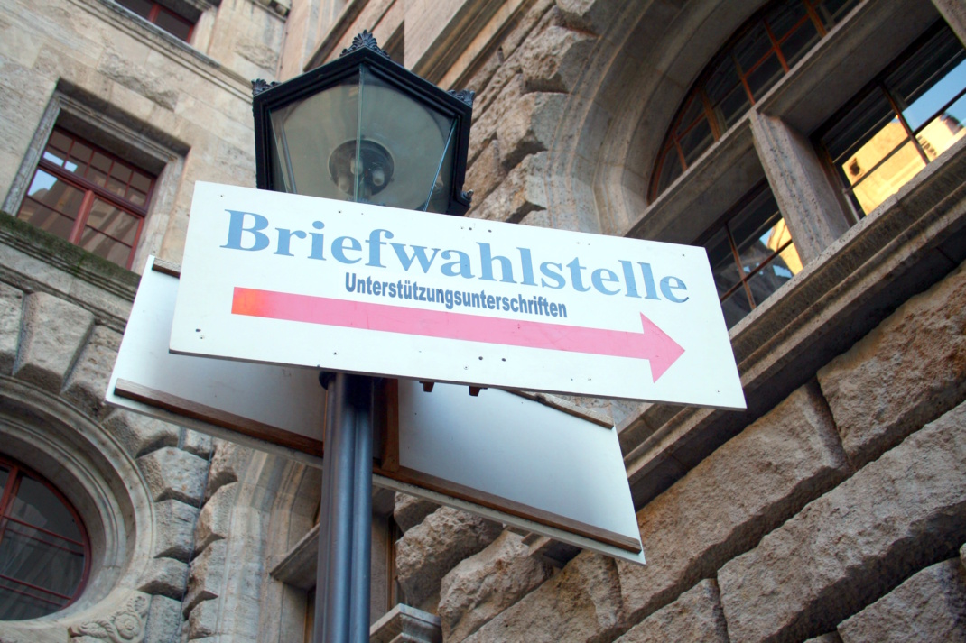 Wegweiser zur Briefwahlstelle ist an Laterne angebracht, Mauer eines Gebäudes im Hintergrund.