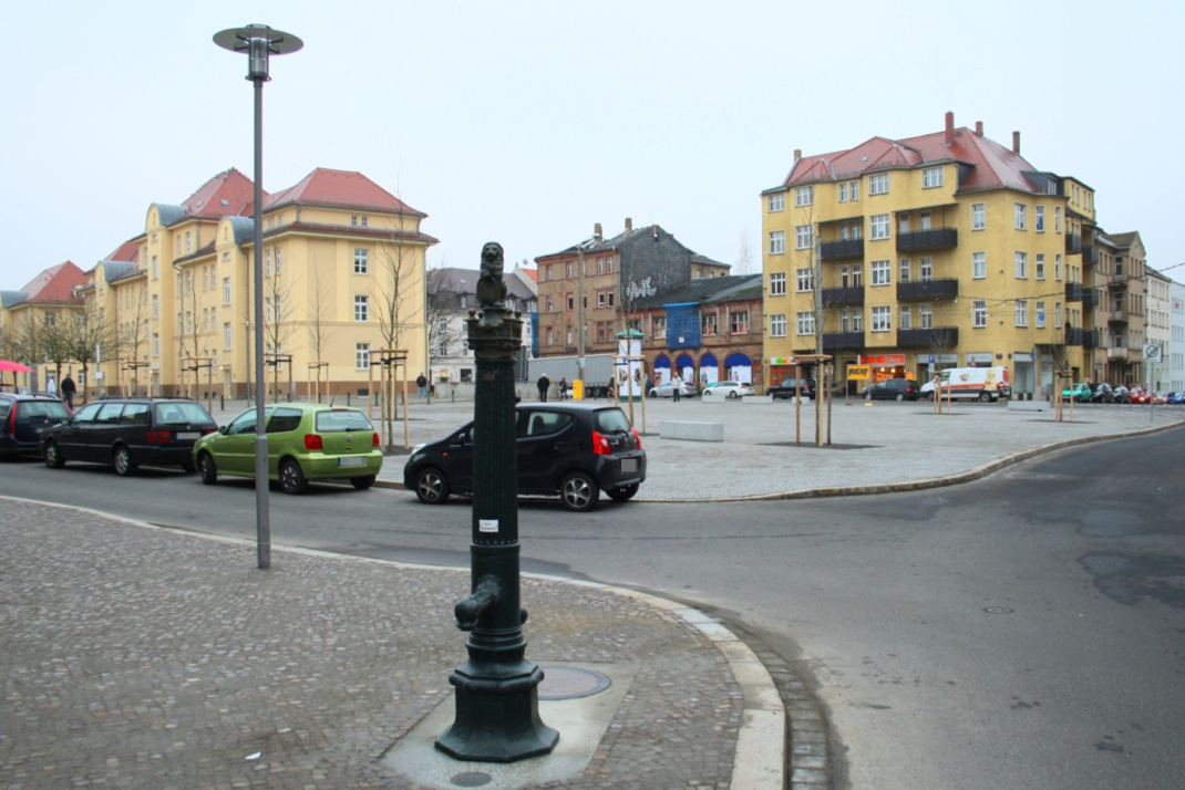 Freier Platz mit Häusern, Wasserpumpe und parkende Kfz,