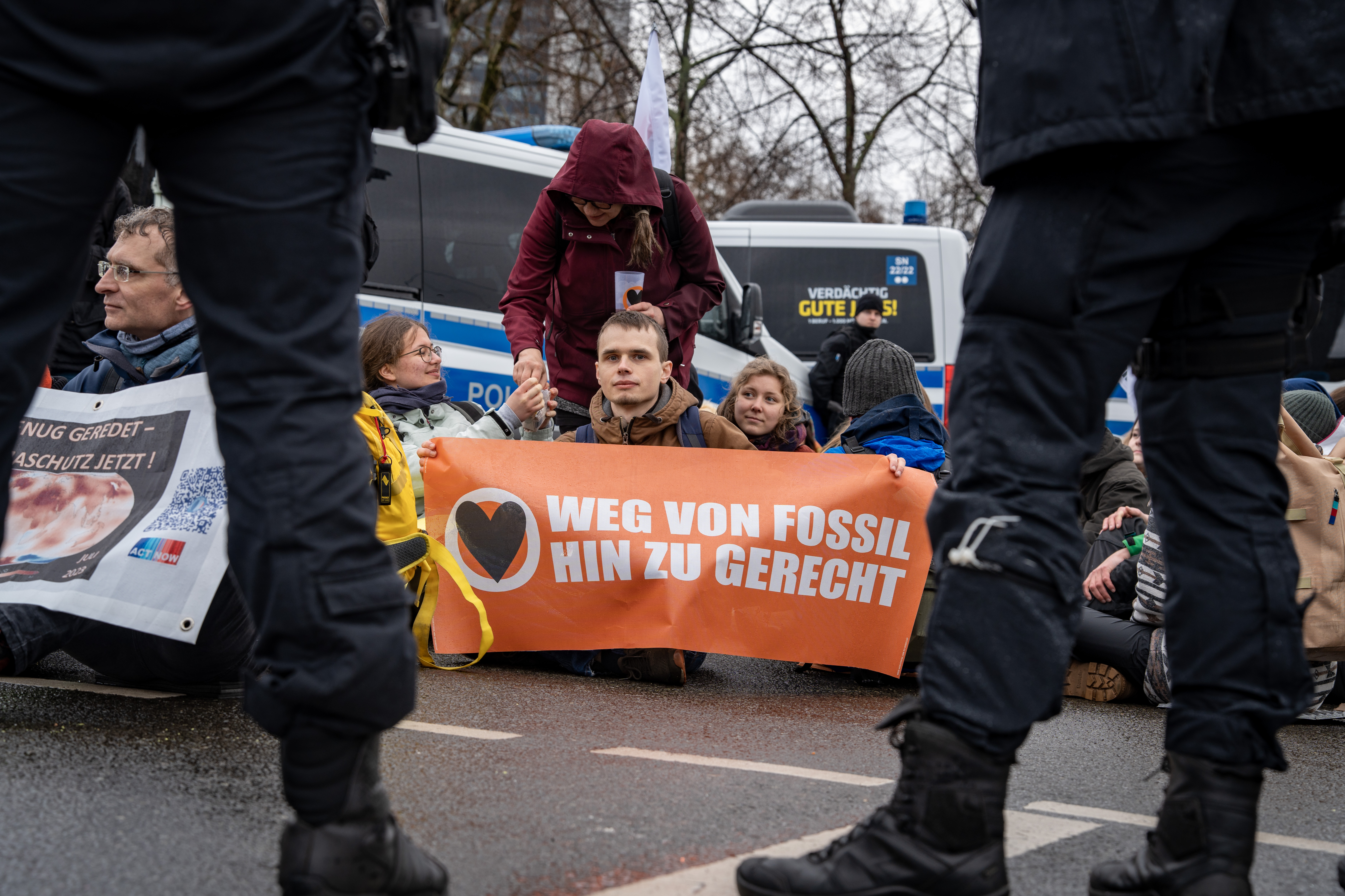 Demonstranten sitzen hinter Transparent, umgeben von Polizeikräften.