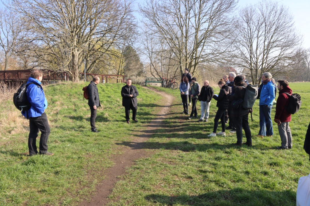 Mehrere Personen stehen an einem Pfad, Wiese, Bäume und blauer Himmel sind zu sehen.
