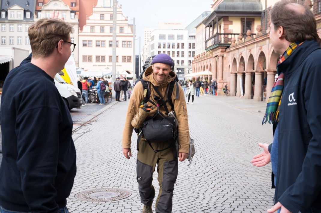 Mann mit Rucksack wird von zwei anderen enpfangen.