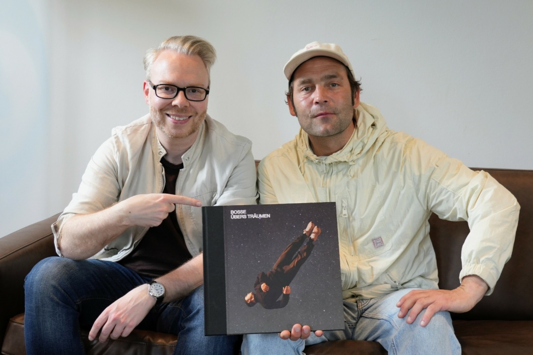 Zwei männliche Personen schauen sitzend zur Kamera und halten ein Buchcover.