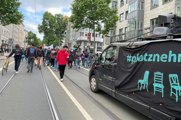 Menschenmenge läuft eine Straße entlang, im Vordergrund ist ein Auto zu sehen.