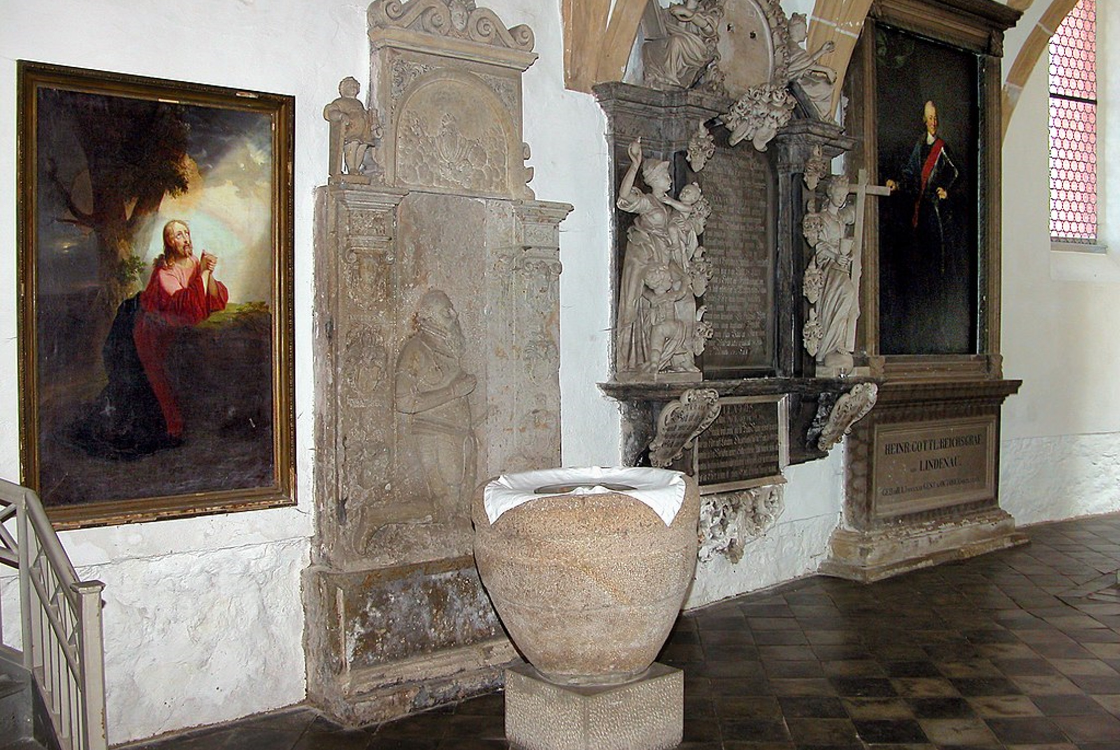 Taufstein vor Epitaphen und Gemälden (Jörg Blobelt, CC BY-SA 3.0, https://commons.wikimedia.org/wiki/Category:Interior_of_St._Nikolai_(Machern)?uselang=de#/media/File:20090425285DR_Machern_Nikolaikirche_im_Chorraum.jpg)