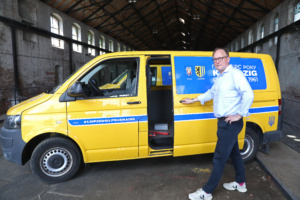 Mann steht vor blau-gelbem Transportwagen.