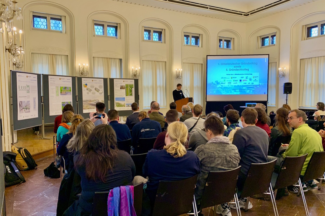 Menschen sitzen bei Podium vor einem Monitor und hören einen Vortrag.