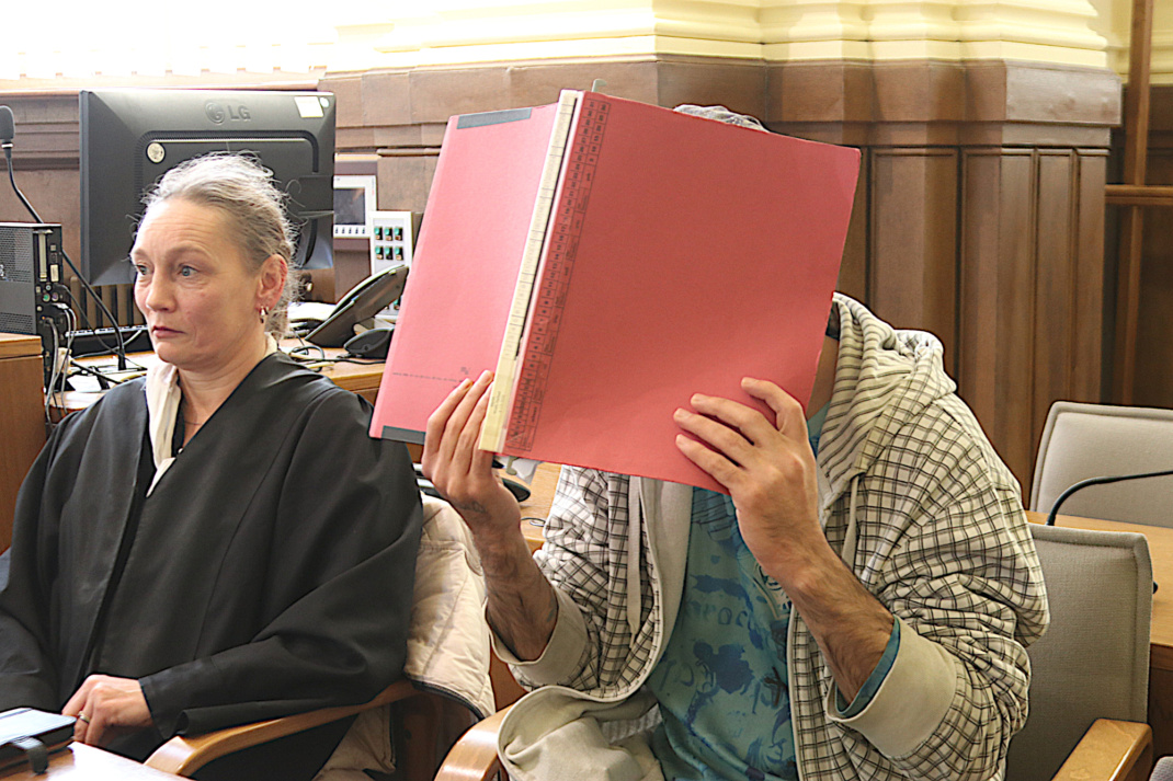 Angeklagter, der sein Gesicht hinter einem roten Ordner verbirgt, mit seiner Verteidigerin vor Gericht.