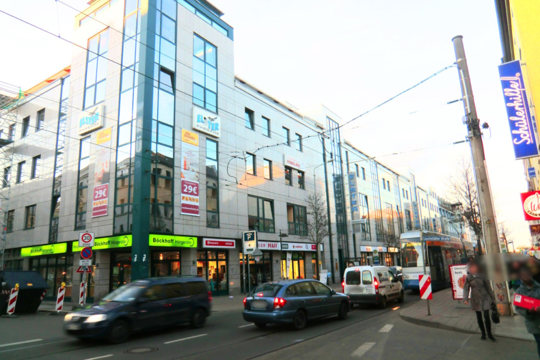 Stark befahrene Straße an Einkaufszentrum, Autos, Passanten auf Gehsteig.