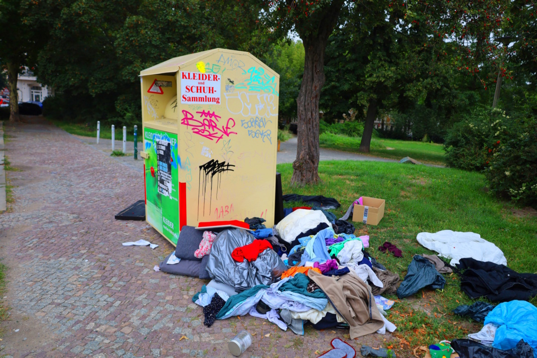 Altkleider-Container, aufgehäufte Textilien, dahinter Park und Wiese.